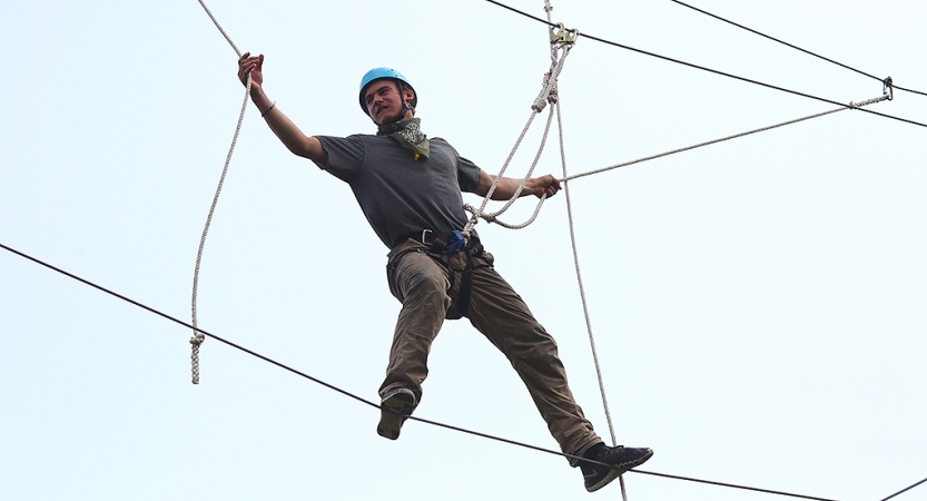 A person wearing safety gear and secured by ropes balances on an obstacle on a high ropes course. 